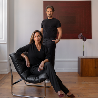 Andre Kvamme and Vanessa Lindblad pose in a minimalist room with a large red abstract painting on the wall. Vanessa sits in a black leather chair, and the man stands behind her wearing a black t-shirt and black pants.