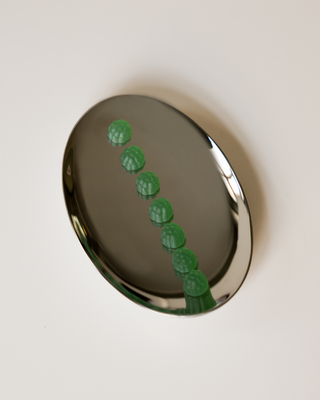 A polished silver oval tray containing seven small, textured green supplements arranged in a diagonal line, set against a solid off-white background.