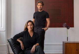 Andre Kvamme and Vanessa Lindblad pose in a minimalist room with a large red abstract painting on the wall. Vanessa sits in a black leather chair, and the man stands behind her wearing a black t-shirt and black pants.