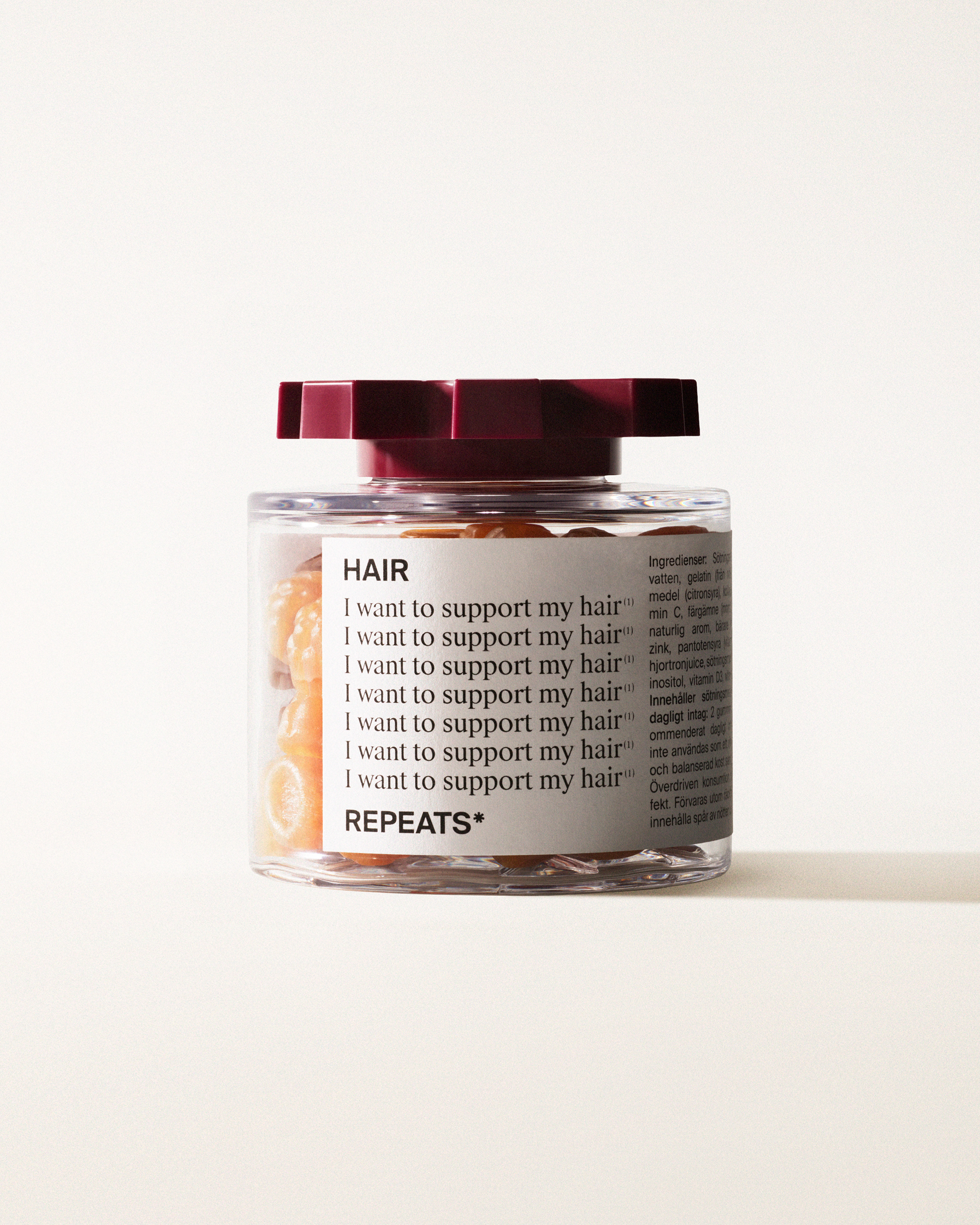 A transparent jar of REPEATS Hair, filled with orange gummies (supplements), sealed with a dark red, shaped lid. The jar has a white label featuring the text "Hair", repeated rows of "I want to support my hair", and the brand Logo