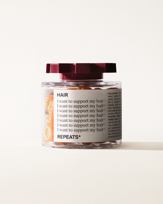 A transparent jar of REPEATS Hair, filled with orange gummies (supplements), sealed with a dark red, shaped lid. The jar has a white label featuring the text "Hair", repeated rows of "I want to support my hair", and the brand Logo