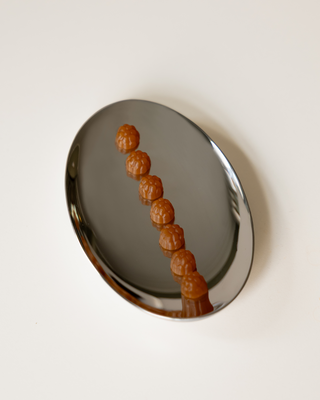 A polished silver oval tray containing seven small, textured orange supplements arranged in a diagonal line, set against a solid off-white background.