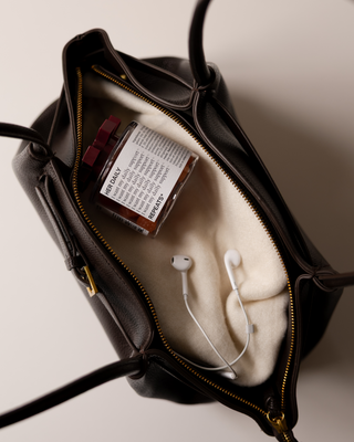 A dark brown leather handbag shown open, revealing a cream-colored interior lining. Inside, a small repeats jar labeled "Her Daily" and white wired earbuds are placed on a neutral off-white background.