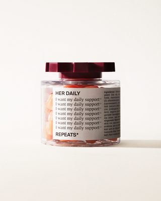 A transparent jar of REPEATS Her Daily, filled with pink gummies (supplements), sealed with a dark red, shaped lid. The jar has a white label featuring the text "Her Daily", repeated rows of "I want my daily support", and the brand Logo