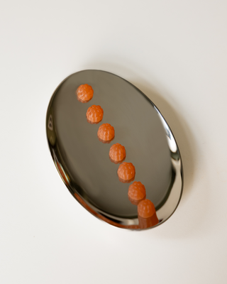 A polished silver oval tray containing seven small, textured orange supplements arranged in a diagonal line, set against a solid off-white background.