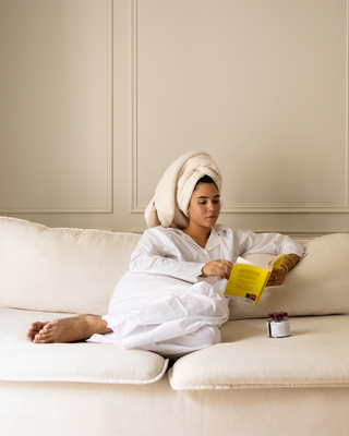 A woman relaxes on a cream-colored sofa wearing a white robe and a white towel wrapped around her head. She is holding a REPEATS product, unscrewing the lid. 