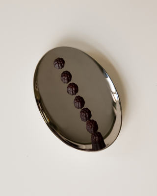 A polished silver oval tray containing seven small, textured dark-purple supplements arranged in a diagonal line, set against a solid off-white background.