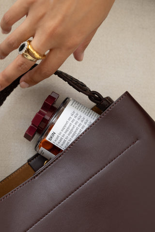 A close-up of a dark brown leather shoulder bag with a braided strap, shown holding a REPEATS bottle. A person