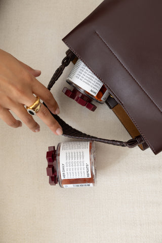 A dark brown leather shoulder bag with a braided strap, shown on a textured beige fabric background next to a small REPEATS jar with a white label and burgundy cap.
