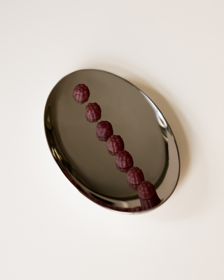 A polished silver oval tray containing seven small, textured purple supplements arranged in a diagonal line, set against a solid off-white background.