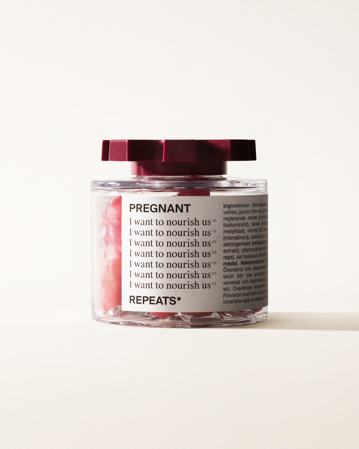 A transparent jar of REPEATS Her Daily, filled with pink gummies (supplements), sealed with a dark red, shaped lid. The jar has a white label featuring the text "Pregnant", repeated rows of "I want to nourish us", and the brand Logo
