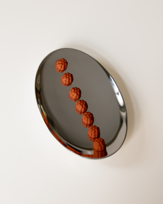 A polished silver oval tray containing seven small, textured orange supplements arranged in a diagonal line, set against a solid off-white background.