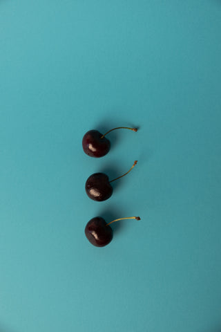 Three dark red cherries with long stems arranged vertically on a solid turquoise blue background.