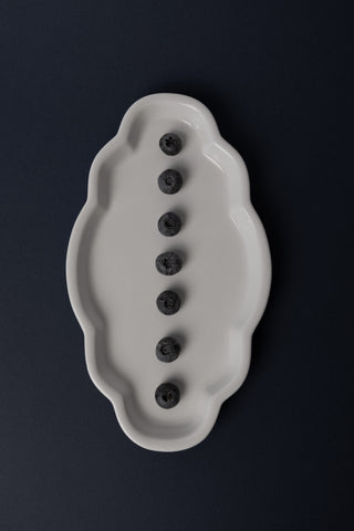 A white ceramic dish with an irregular scalloped border, containing seven dark blueberries arranged in a vertical line, set against a solid dark blue background.
