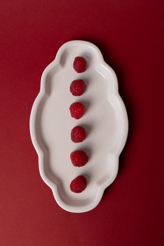 A white ceramic dish with scalloped edges holds six red raspberries arranged vertically, set against a solid dark red background.