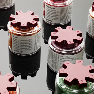 Close-up of several REPEATS jars with burgundy lids and product visible through clear tops, arranged on a reflective surface. One label clearly reads REPEATS and HAIR.